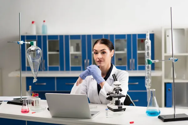 Scientist in latex gloves looking at camera near medical equipment and laptop in lab — Stock Photo