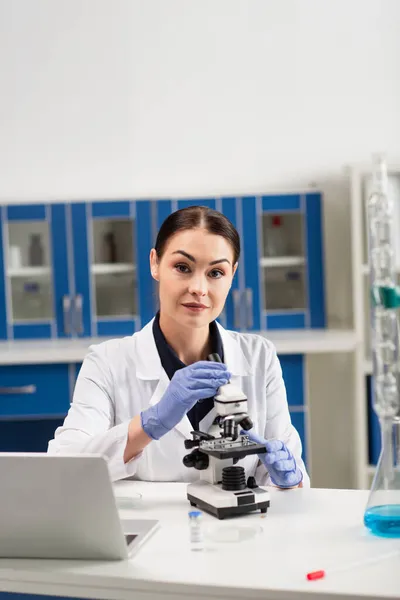 Morena cientista em luvas de látex olhando para a câmera perto de microscópio e laptop em laboratório — Fotografia de Stock