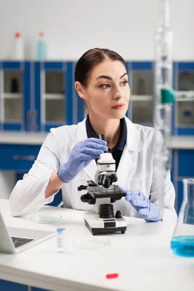 Scienziato in mantello bianco con microscopio vicino a fiaschetta e laptop in laboratorio — Foto stock