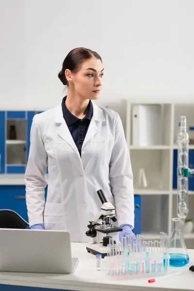 Scientifique en blouse blanche debout à proximité de matériel médical, ordinateur portable et vaccin en laboratoire — Photo de stock