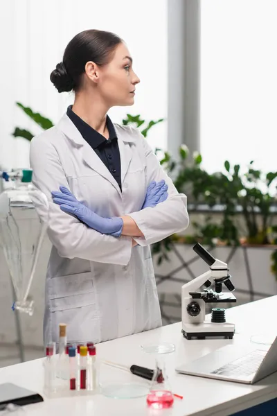 Vista lateral de científico morena en bata blanca de pie cerca del microscopio y tubos de ensayo en laboratorio - foto de stock