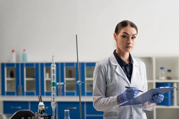 Scientist holding clipboard and looking at camera near equipment in lab — Stock Photo