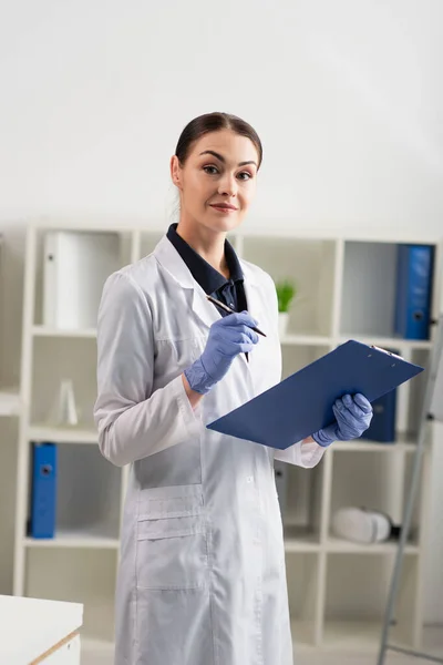 Scientifique en manteau blanc regardant la caméra tout en tenant le stylo et le presse-papiers dans le laboratoire — Photo de stock