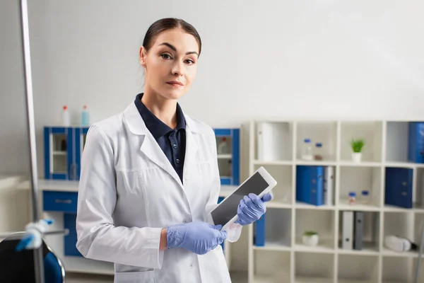 Cientista em luvas de látex olhando para a câmera e segurando tablet digital no laboratório — Fotografia de Stock