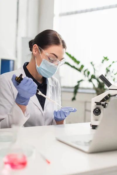 Científico en guantes de látex y máscara médica que trabaja con placa Petri y pipeta electrónica en laboratorio - foto de stock