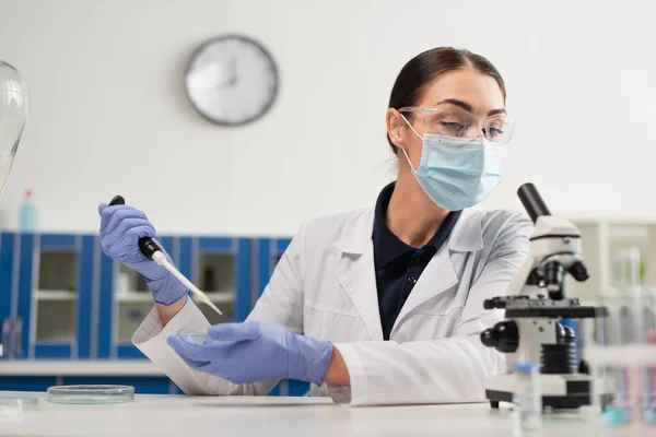 Cientista em óculos e luvas de látex trabalhando com pipeta eletrônica e microscópio em laboratório — Fotografia de Stock