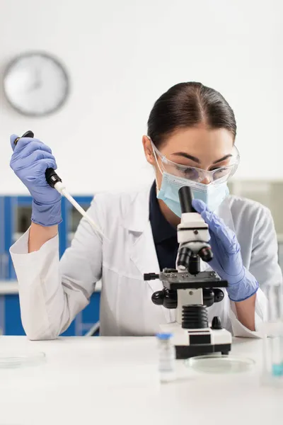 Scientifique en masque médical et gants en latex tenant une pipette électronique près du microscope et des boîtes de Pétri en laboratoire — Photo de stock