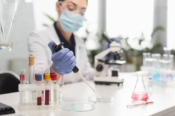 Científico borroso en guante de látex trabajando con pipeta electrónica y placas de Petri cerca de tubos de ensayo con muestras de sangre en laboratorio — Stock Photo