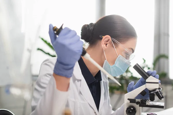 Side view of scientist in goggles and medical mask holding blurred pipette and using microscope in lab — Stock Photo