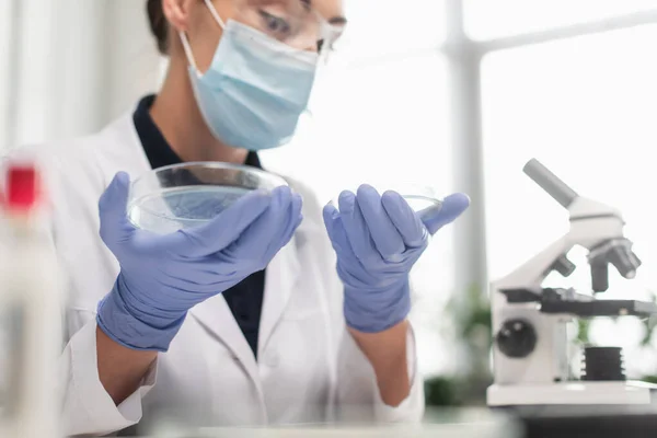 Científico borroso en guantes de látex y máscara médica que sostiene placas de Petri cerca del microscopio en el laboratorio - foto de stock