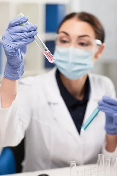 Verschwommener Wissenschaftler in medizinischer Maske und Latexhandschuhen blickt im Labor auf das Reagenzglas — Stockfoto