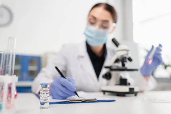 Коронавирусная вакцина рядом с размытым ученым, пишущим на планшете возле микроскопа в лаборатории — стоковое фото