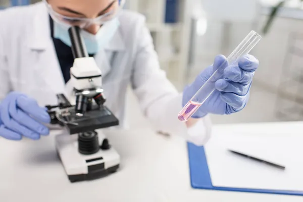 Tubo de ensaio na mão do cientista desfocado em luvas de látex utilizando microscópio — Fotografia de Stock