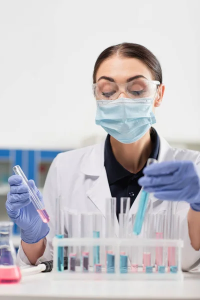 Brünette Wissenschaftlerin in Latexhandschuhen und medizinischer Maske mit Reagenzgläsern in der Nähe von Pipette und Kolben im Labor — Stockfoto