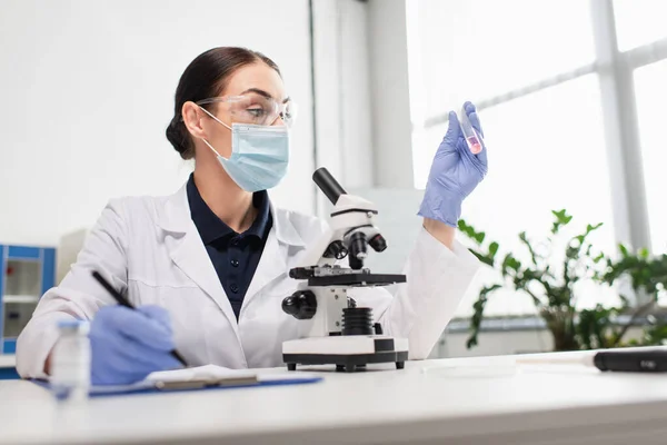 Wissenschaftler in weißem Mantel und Brille mit Reagenzglas und Schrift auf Klemmbrett — Stockfoto