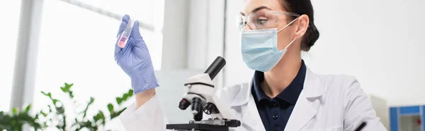 Scientifique en gant de latex et masque médical tenant un tube à essai près du microscope en laboratoire, bannière — Photo de stock
