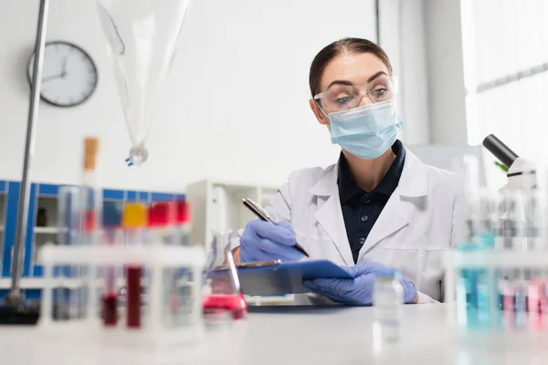 Scientist in goggles writing on clipboard near test tubes and flasks — Stock Photo