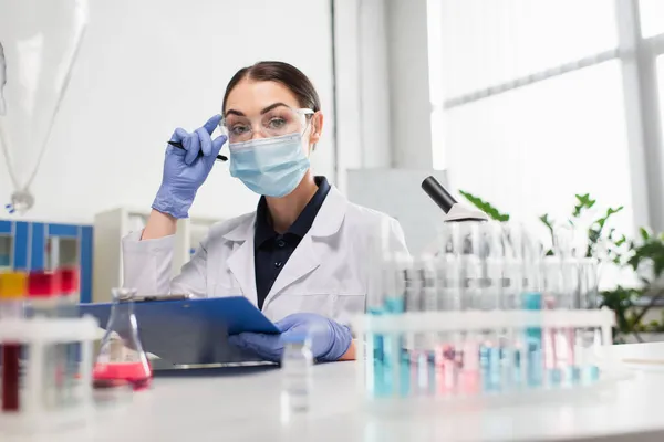 Cientista em óculos segurando prancheta perto de tubos de ensaio e microscópio em laboratório — Fotografia de Stock