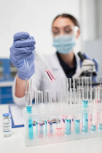 Blurred scientist in latex gloves holding test tube near coronavirus vaccine in lab — Stock Photo