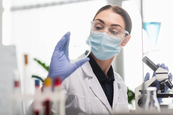 Scientist in latex gloves and medical mask holding petri dish near microscope in lab — Stock Photo