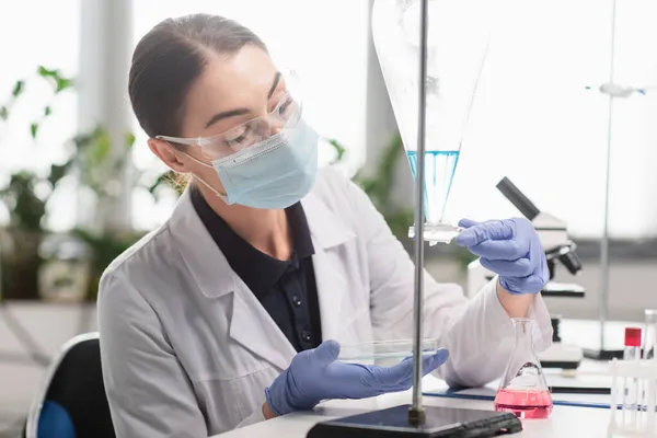 Brünette Wissenschaftlerin mit Brille und medizinischer Maske hält Petrischale in der Nähe von Kolben im Labor — Stockfoto