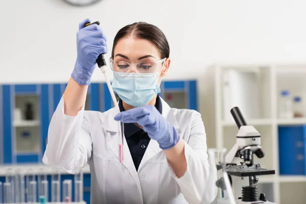 Científico en gafas y máscara médica sosteniendo pipeta electrónica y tubo de ensayo cerca del microscopio - foto de stock