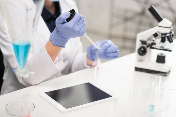 Vista recortada del científico en guantes de látex con pipeta electrónica cerca de tubos de ensayo, microscopio y tableta digital - foto de stock