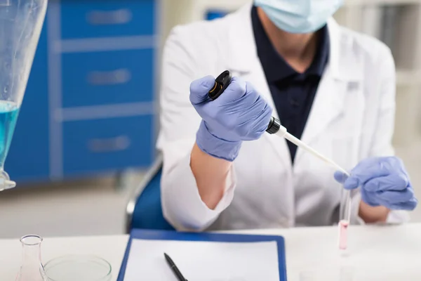 Vue recadrée du scientifique à l'aide d'une pipette électronique tout en travaillant avec un tube à essai près du presse-papiers en laboratoire — Photo de stock