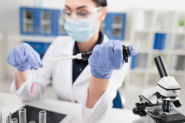 Blurred scientist in medical mask holding electronic pipette near test tubes, microscope and digital tablet — Stock Photo