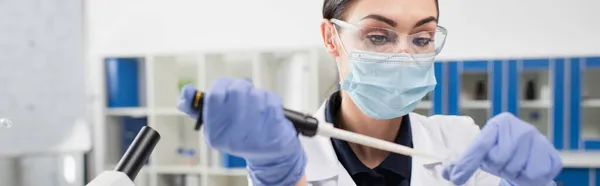 Científico en máscara médica y guantes de látex trabajando con pipeta en laboratorio, pancarta - foto de stock