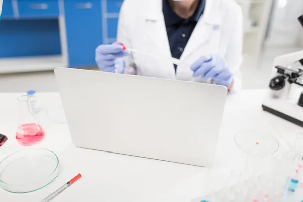Vista cortada do laptop perto de tubos de ensaio e cientista desfocado segurando teste de pcr em laboratório — Fotografia de Stock