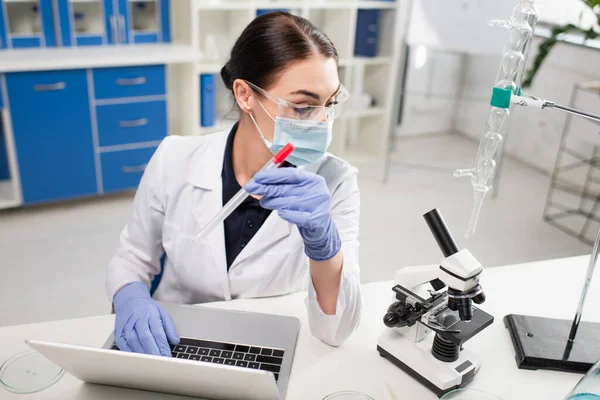 Científico en guantes de látex que sostiene la prueba de pcr y el uso de computadora portátil cerca del microscopio en el laboratorio - foto de stock