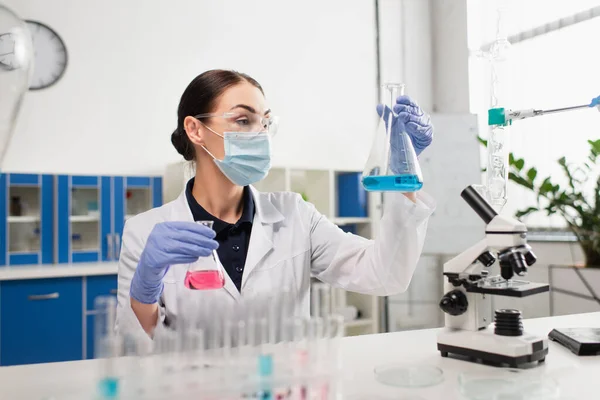 Scientist in safety goggles holding flasks near test tubes and microscope — Stock Photo