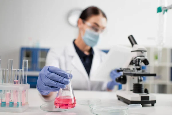 Kolben in der Hand eines verschwommenen Wissenschaftlers mit digitalem Tablet in der Nähe von Petrischalen und Reagenzgläsern — Stockfoto