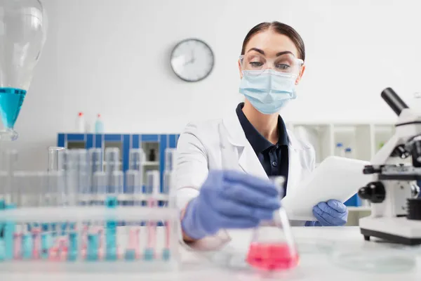 Scientist in goggles holding flask and digital tablet near microscope and blurred test tubes — Stock Photo