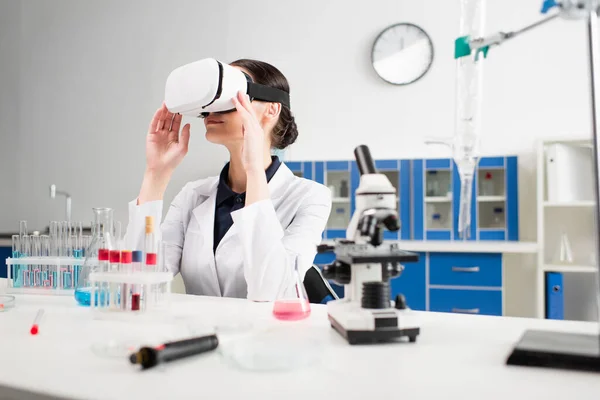 Scientifique en blouse blanche et casque vr près du microscope et des éprouvettes en laboratoire — Photo de stock
