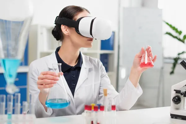 Scientist in virtual reality headset holding flasks near microscope in lab — Stock Photo