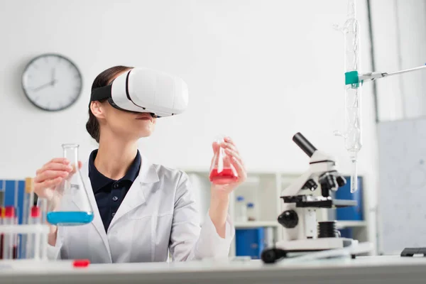 Scientifique en vr casque tenant des flacons tout en travaillant à proximité du microscope et des éprouvettes en laboratoire — Photo de stock