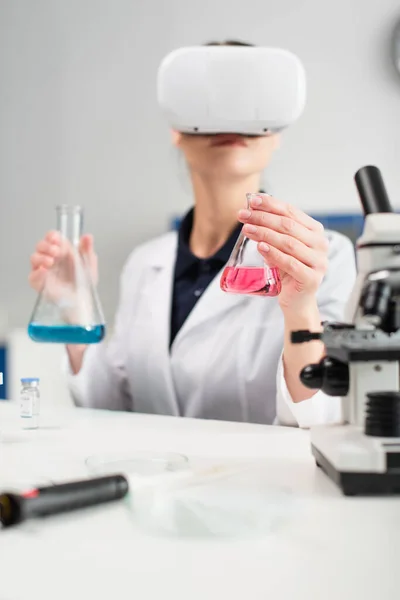 Kolben in der Hand verschwommener Wissenschaftler in der Nähe von Mikroskop, Impfstoff und elektronischer Pipette im Labor — Stockfoto