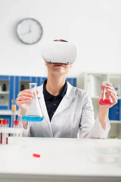 Científico en auriculares de realidad virtual sosteniendo frascos cerca de tubos de ensayo con muestras de sangre en el laboratorio - foto de stock