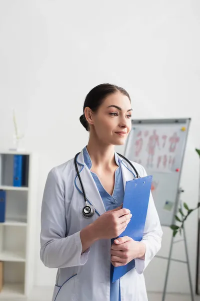 Brunette médecin en manteau blanc et stéthoscope tenant presse-papiers à l'hôpital — Photo de stock