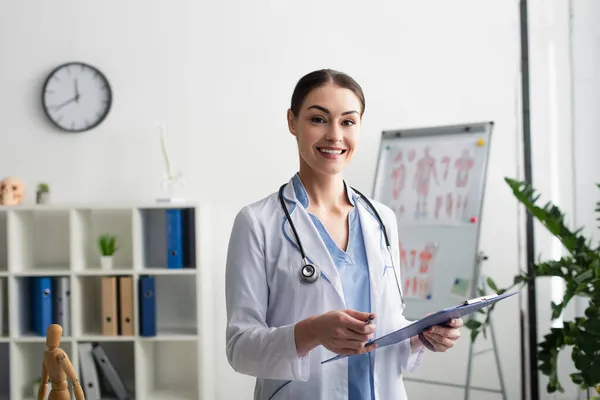 Alegre médico morena sosteniendo pluma y portapapeles en la clínica - foto de stock