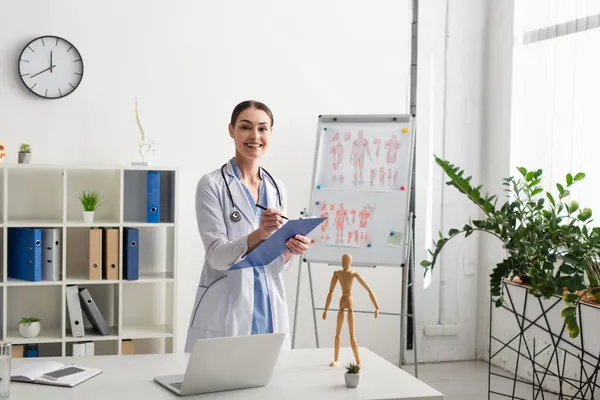 Médico sonriente escribiendo en el portapapeles cerca de dispositivos y agua en la mesa en la clínica - foto de stock