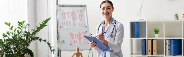 Brunette médecin souriant à la caméra tout en écrivant sur le presse-papiers à l'hôpital, bannière — Photo de stock