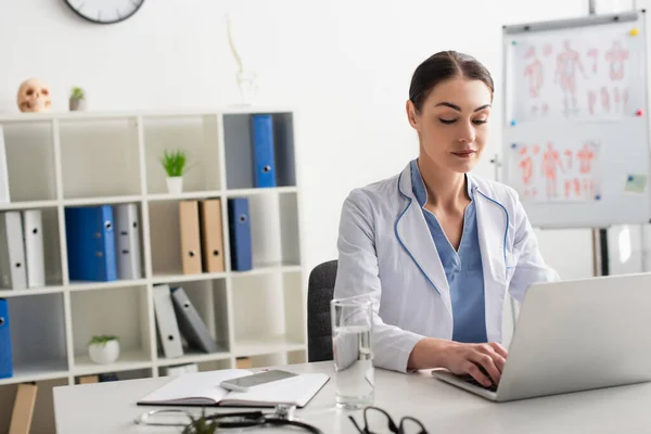 Médico usando laptop perto de notebook e vidro de água na clínica — Fotografia de Stock