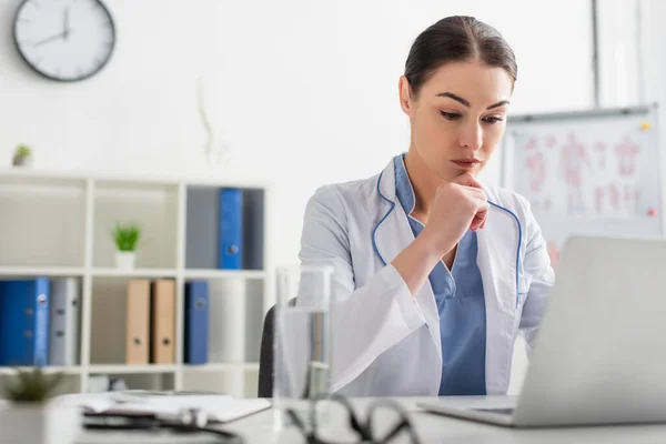 Médico pensativo en bata blanca mirando a la computadora portátil cerca del agua y anteojos en la mesa en la clínica - foto de stock