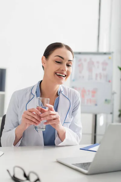 Smiling doctor holding glass of water near laptop and blurred eyeglasses in clinic — Stock Photo