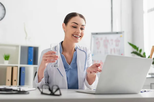 Médecin souriant tenant un verre d'eau lors d'un appel vidéo sur un ordinateur portable à l'hôpital — Photo de stock