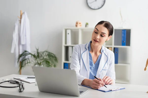 Doctor writing on clipboard and looking at laptop near eyeglasses and stethoscope in clinic — Stock Photo