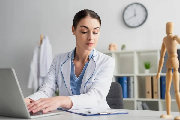 Brunette doctor in white coat using laptop near clipboard in hospital — Stock Photo